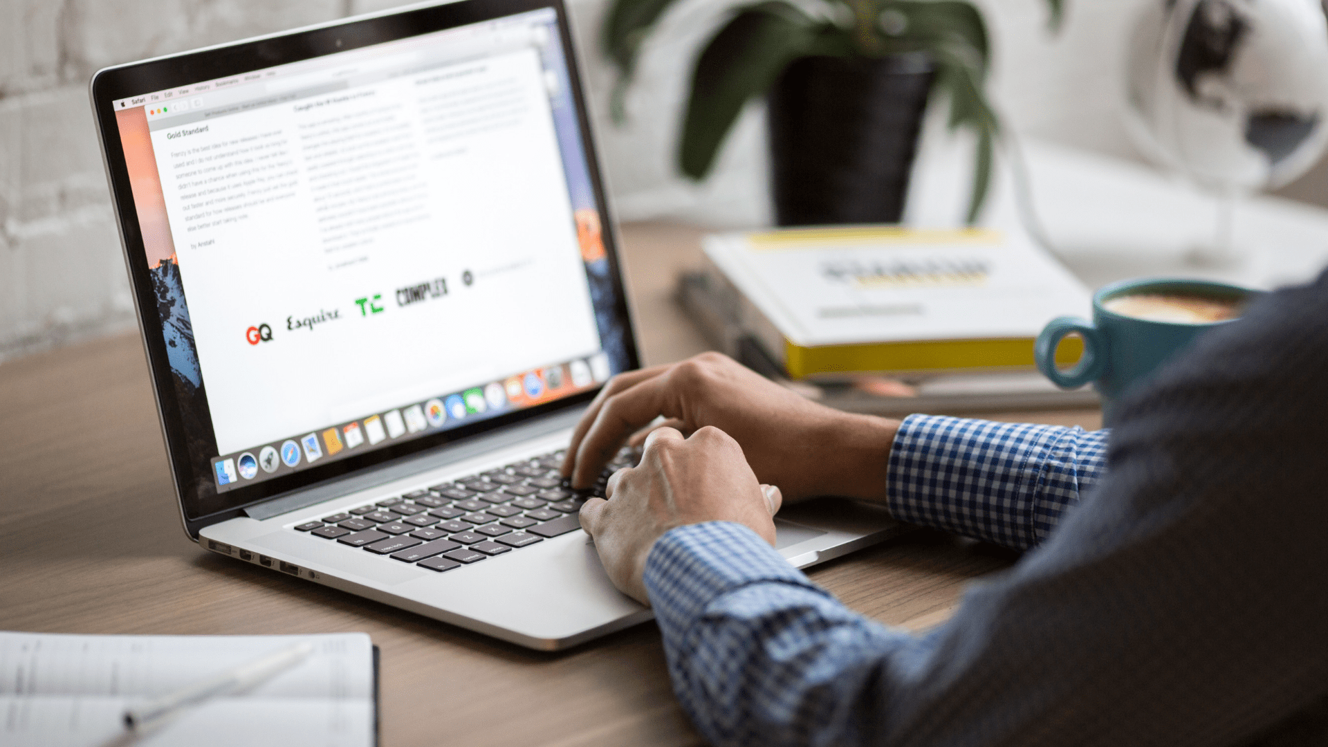 person sits at wood desk on laptop with black keys, reading a blog page, teal cappuccino mug next to them
