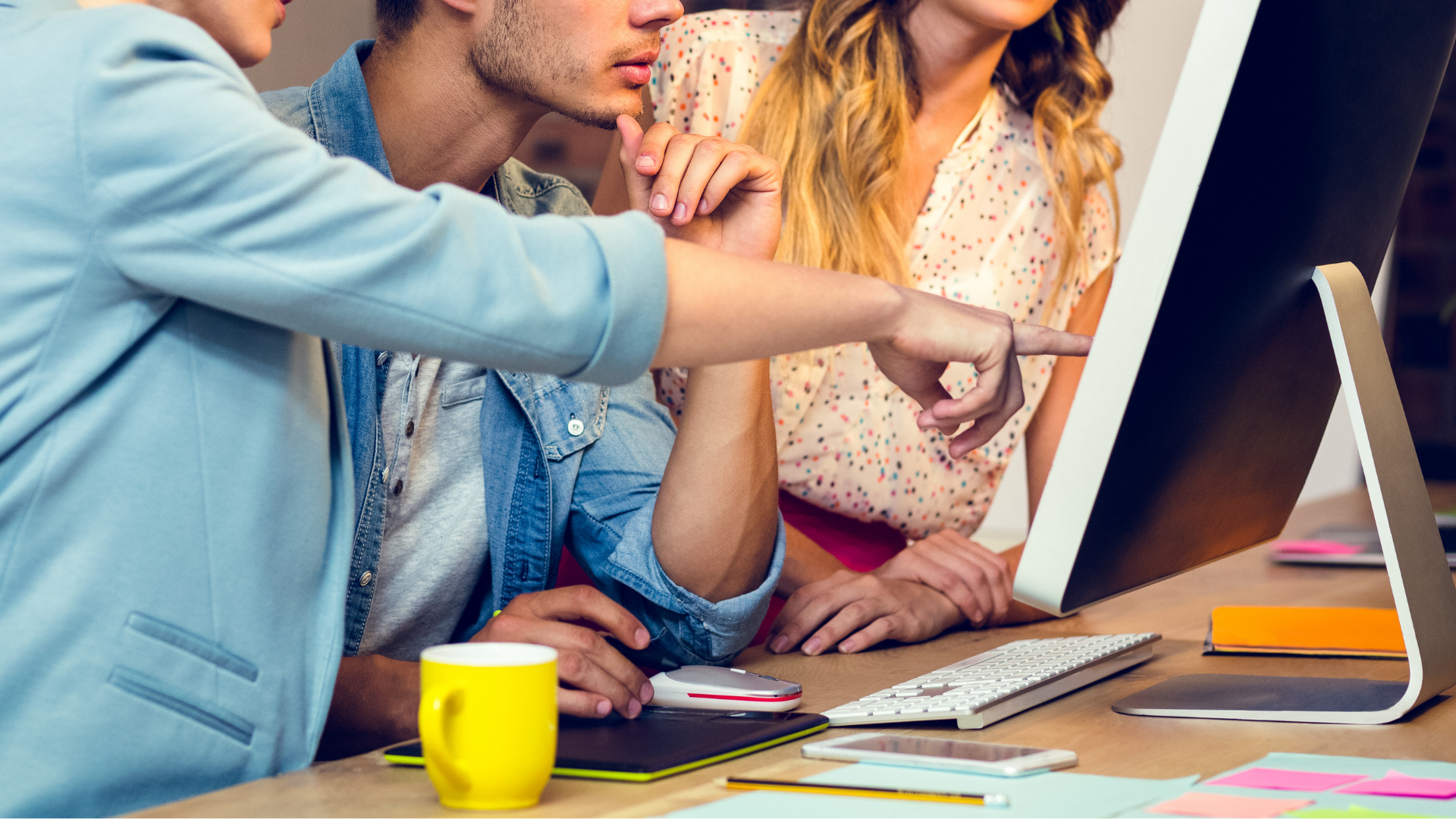 three casually dressed professionals look at a desktop screen, one points at content
