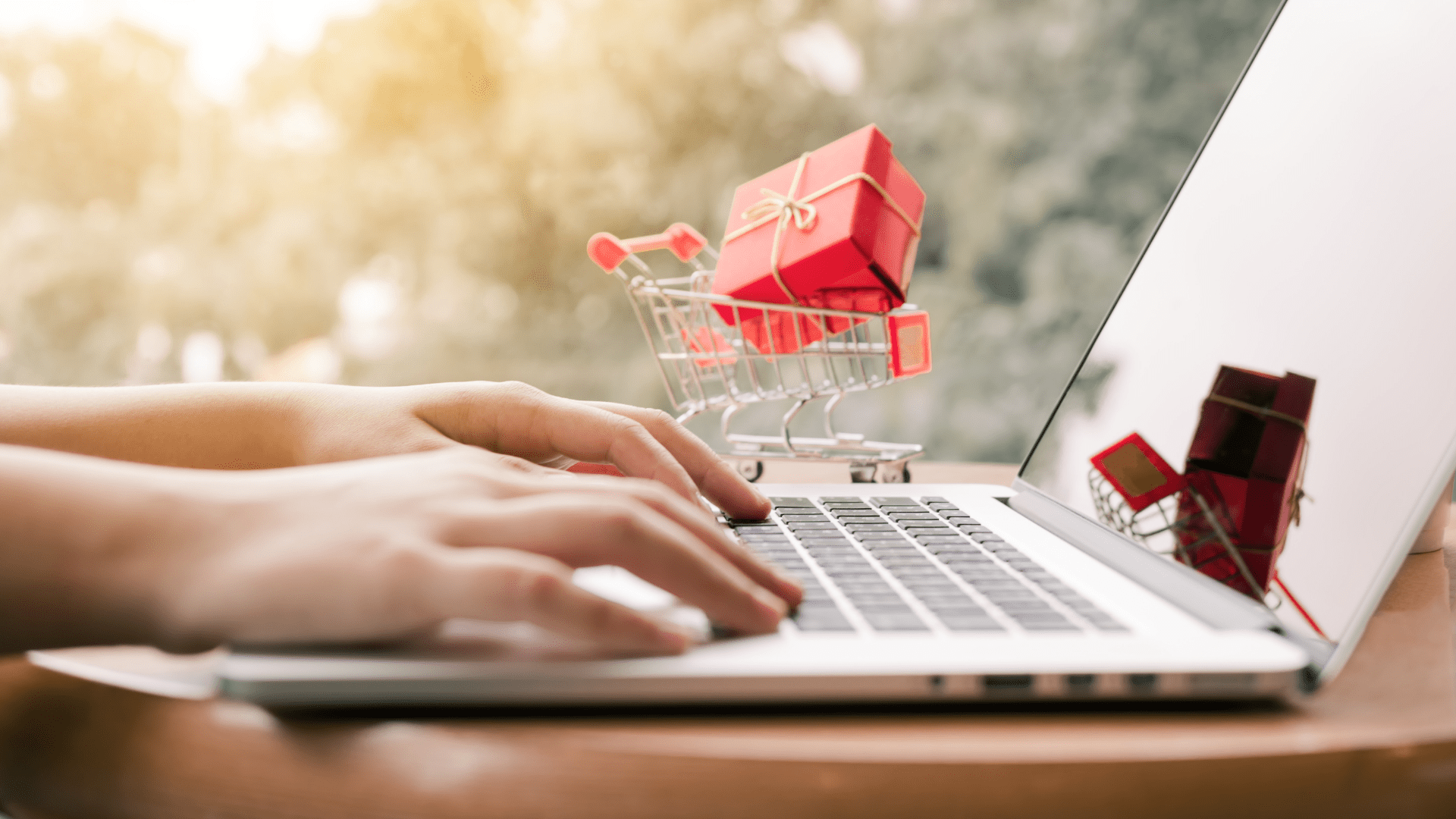 person on a laptop in front of a sunny window with a tiny metal shopping cart with a large scale red package with gold bow