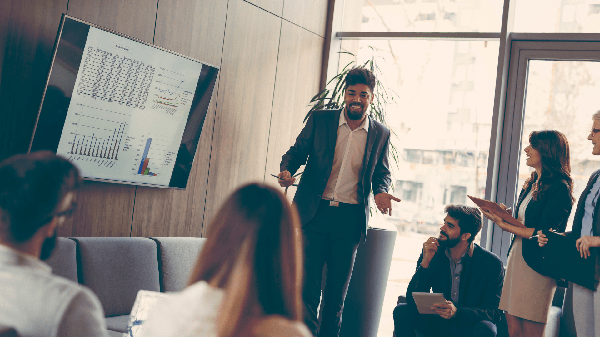 a man in a relaxed business suit with a goatee presents on a monitor to a group of happy people