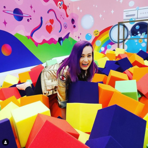 Molly Burke is looking up and laughing at the camera. She is so happy as she is in the middle of colorful foam blocks that are in a very fun foam pit in the Instagram lounge