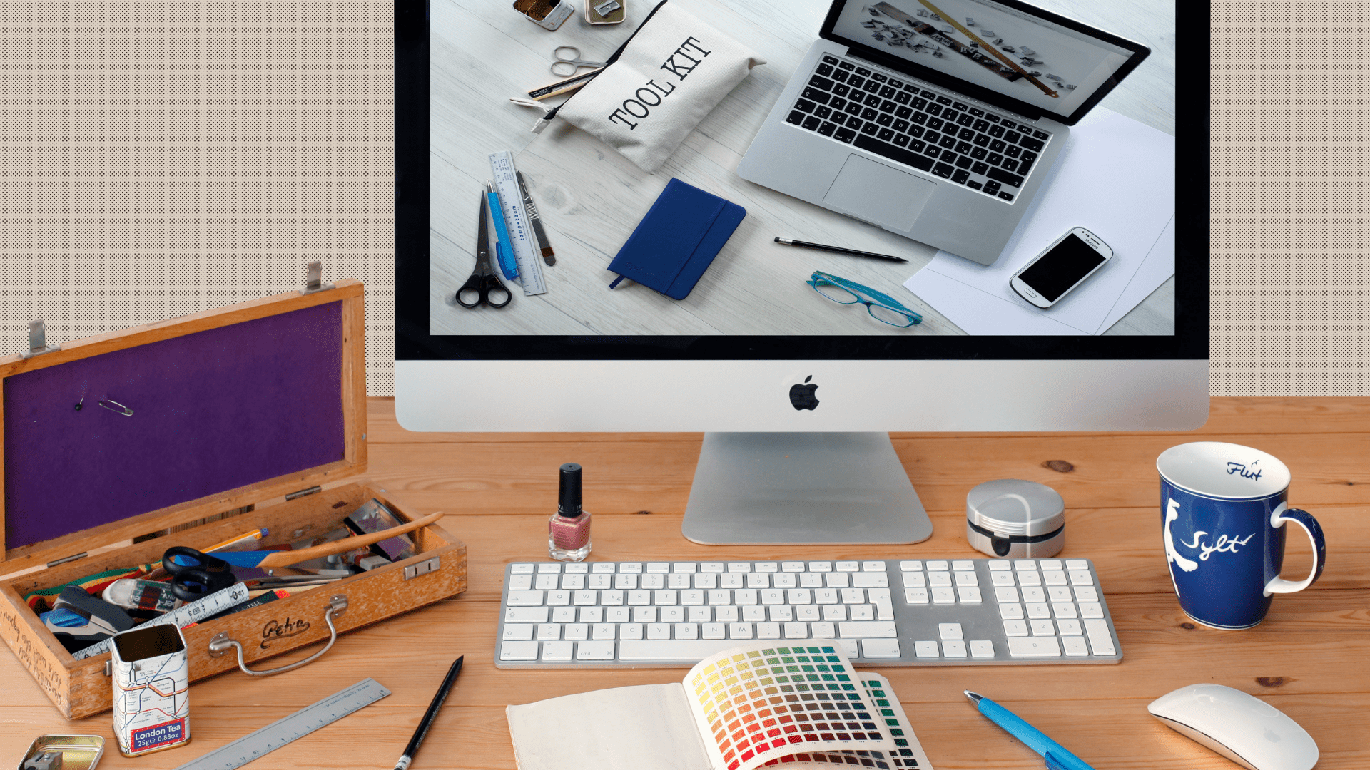 wooden desk with iMac and magic mouse and keyboard has color samples and other design tools scattered about, an image of a toolkit laptop and smartphone is on the screen
